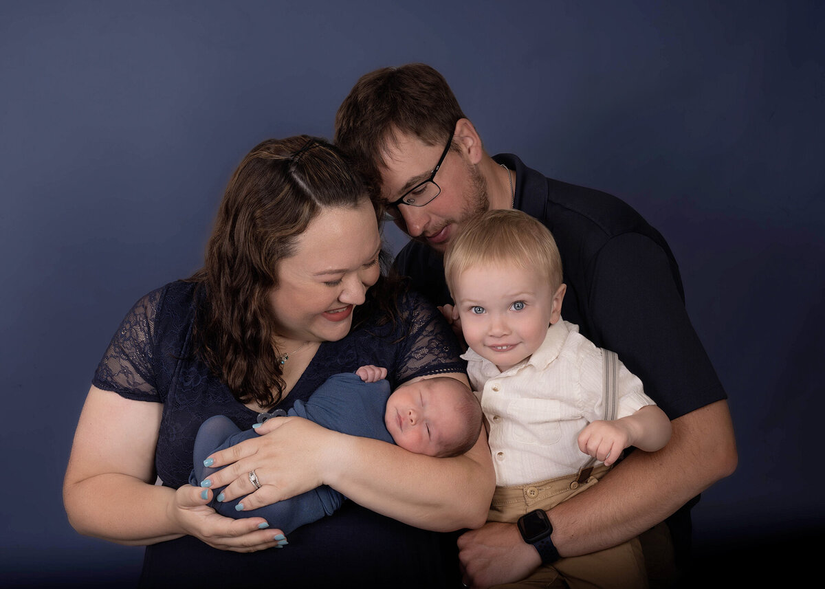 A happy toddler smiles big while in dad's arms and hugging his newborn brother in mom's arms