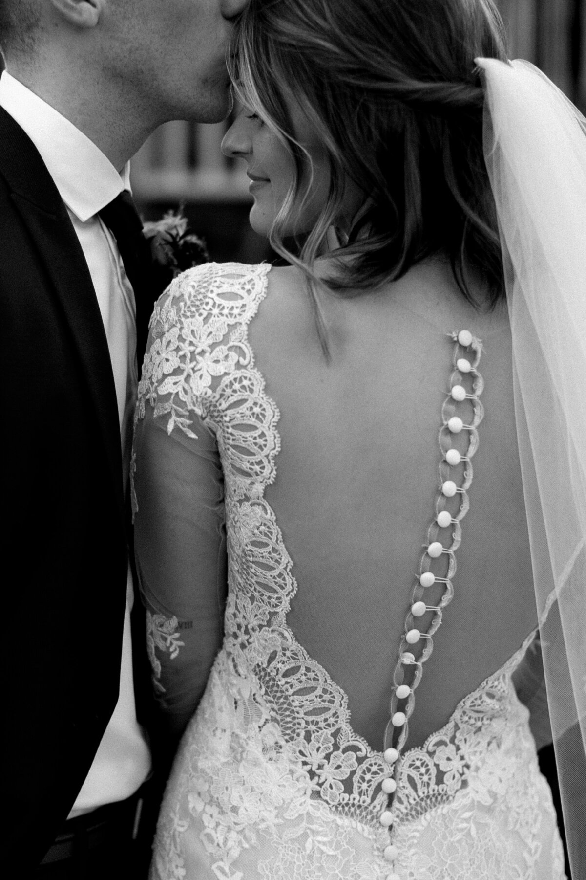Back of brides dress with buttons going down the back and her head turned towards her groom and her groom kissing her on the forehead.