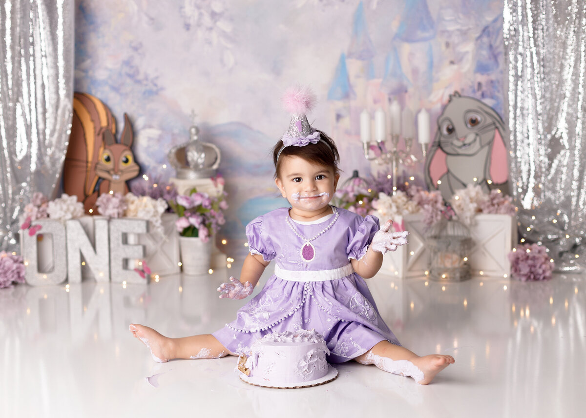 Sofia the First themed cake smash  at West Palm Beach and Delray Beach photography studio. Baby girl is wearing a Sofia the First dress with cake on her legs, hands, and face. She is smiling cheekily at the camera. In the background, there is a purple and blue princess backdrop and flowers.