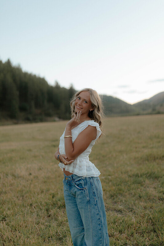 Gently touching her face with one hand, a girl smiles at her senior photos photographer for a posed portrait outside.