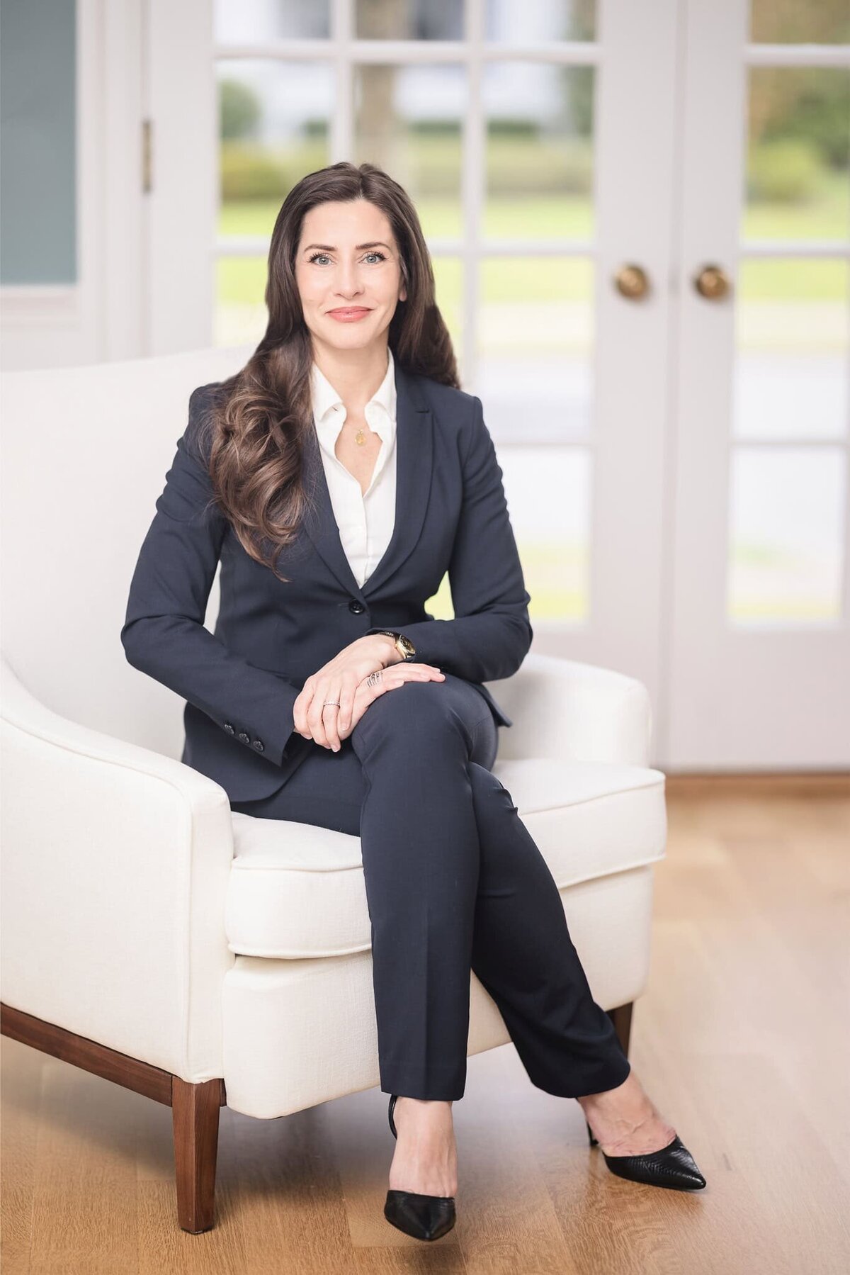 Corporate headshot of a professional woman in a navy suit seated in a white chair, smiling with natural light from French doors, Long Island headshots.