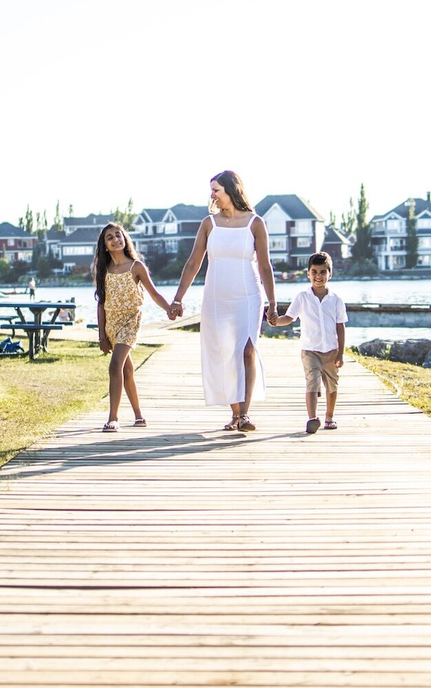 A mom in a white dress is walking near a lake holding her son and daughter's hands