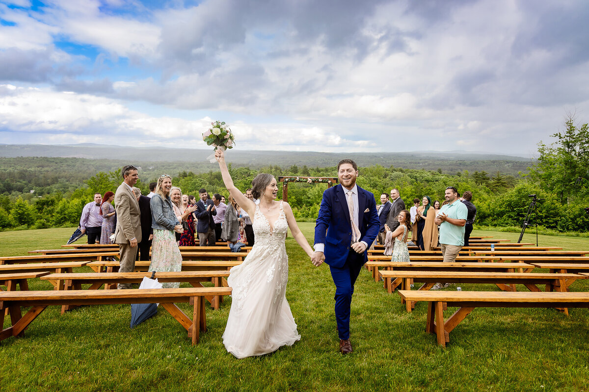 Bride cheering holding hands with groom as they walk back down the aisle after wedding ceremony at Mountain View Stables in Loudon