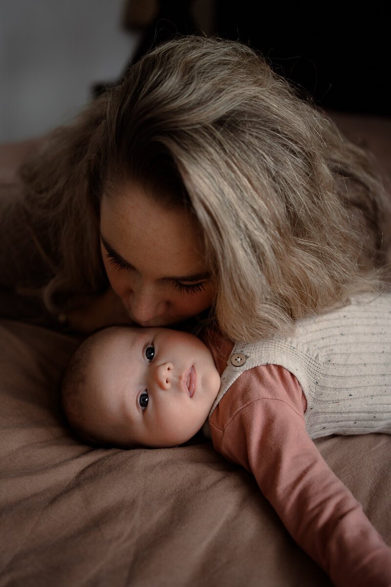 baby ligt op het bed terwijl moeder haar een kusje op haar slaap geef.