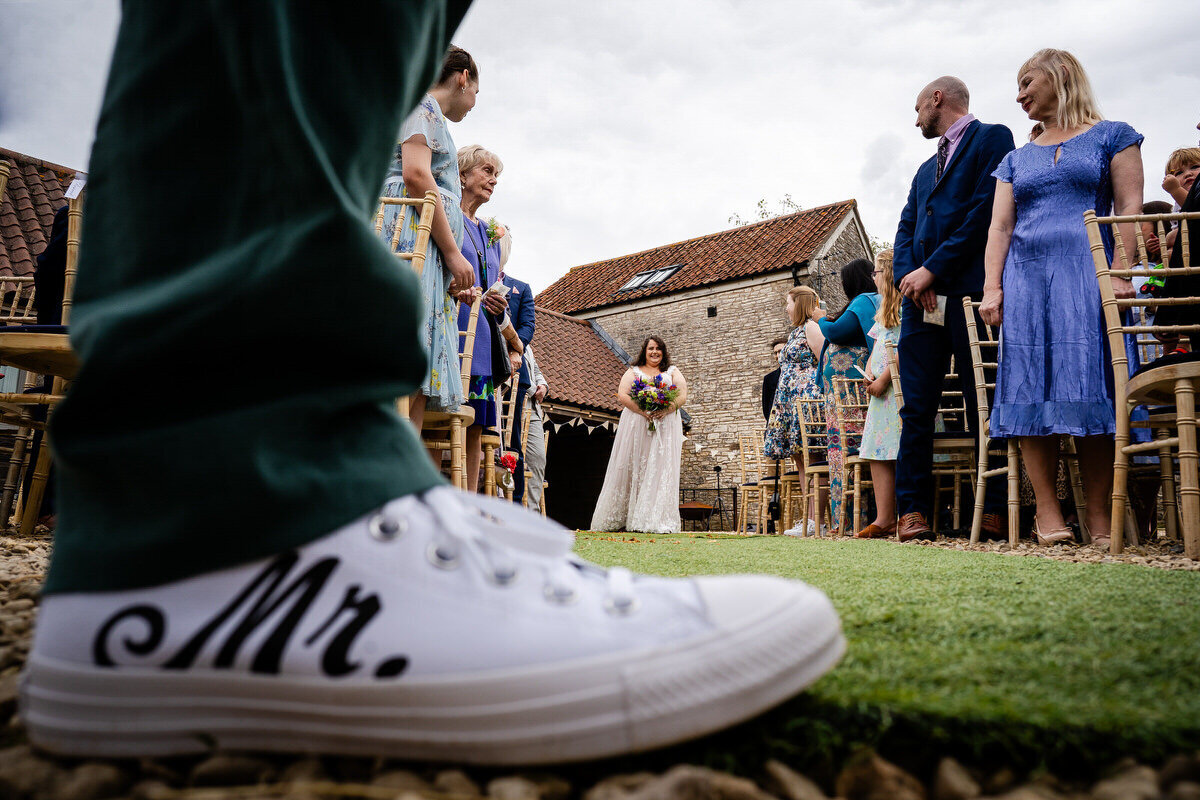 closure up of the grooms personalised "mr" converse shoes in the foreground with the bride in the background as she is walking down the aisle at Folly Farm