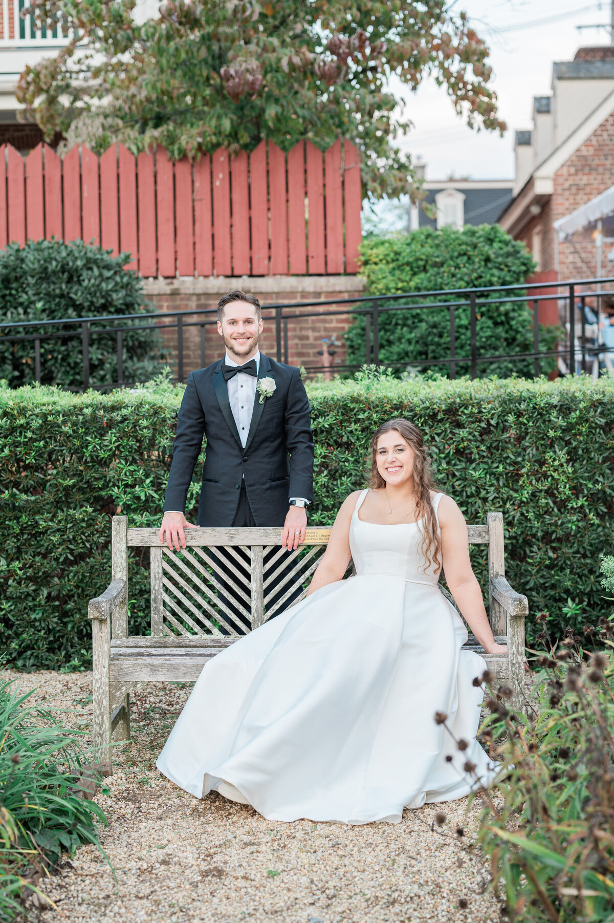 A bride sitting on a bench with a groom standing behind it