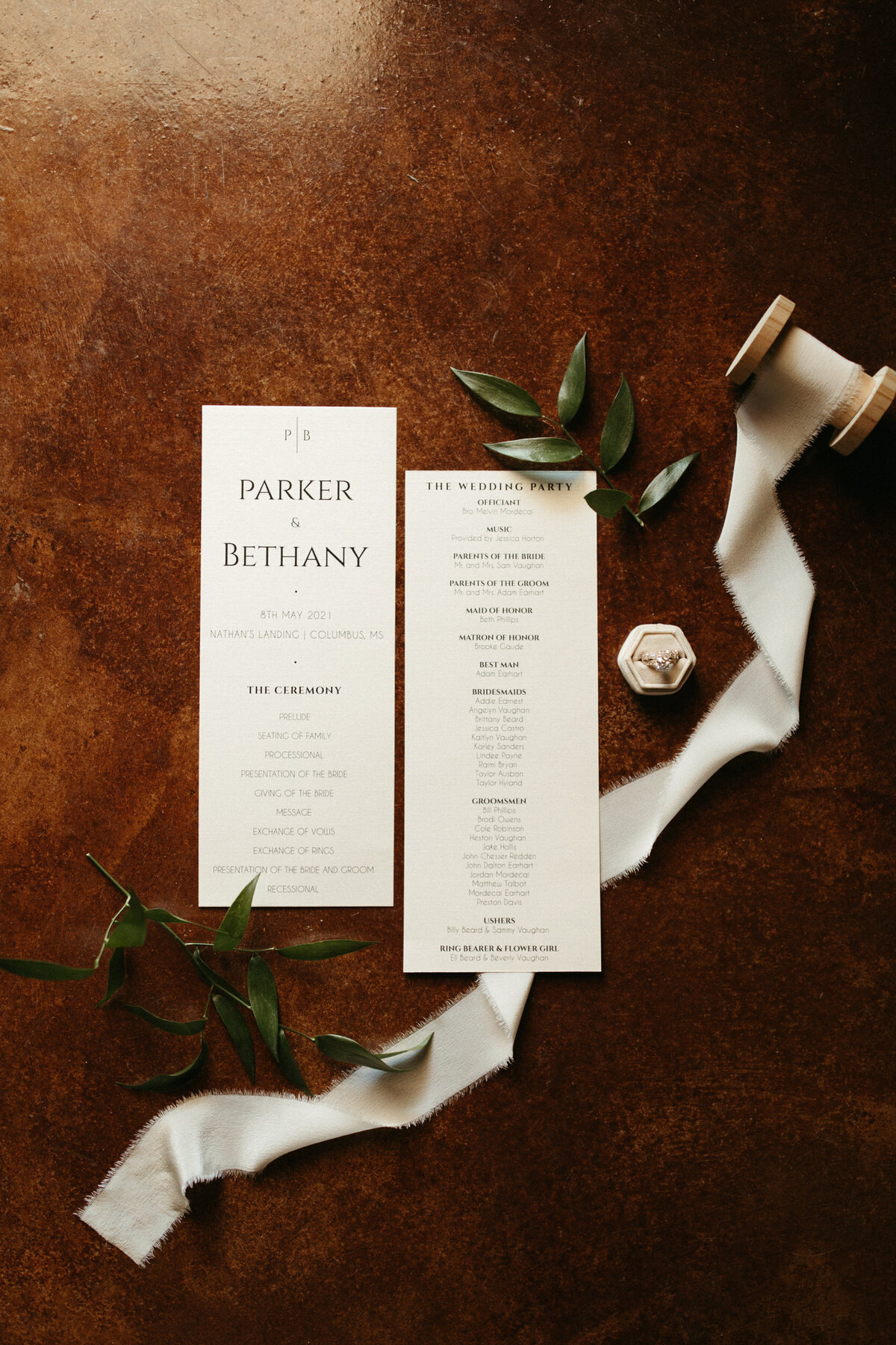 Wedding details flat lay of programs, chiffon ribbon on a spool, greenery, and the ring in a ring box