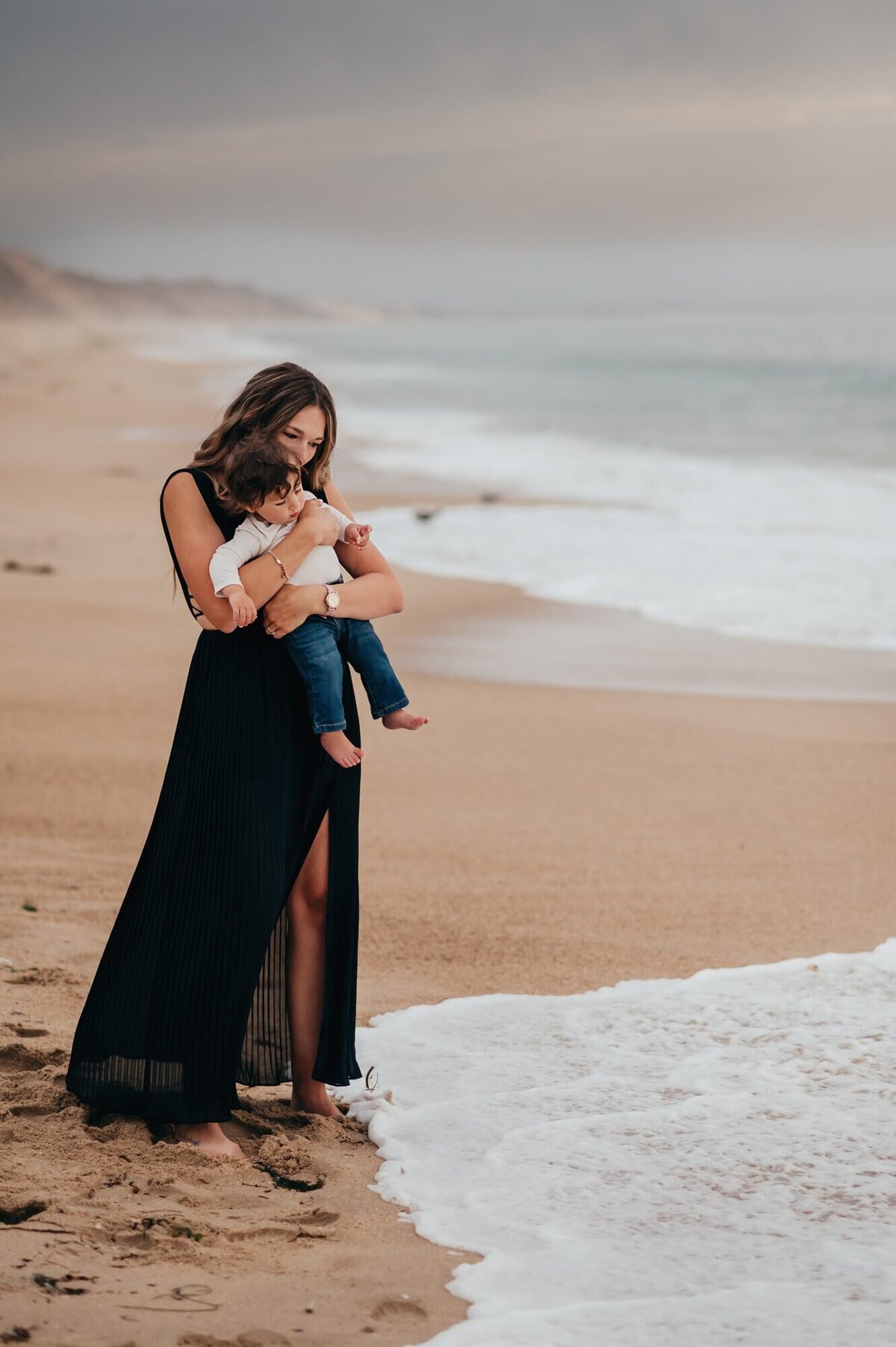 Mother in a long black dress holding her toddler at the edge of the surf on a Monterey beach at dusk