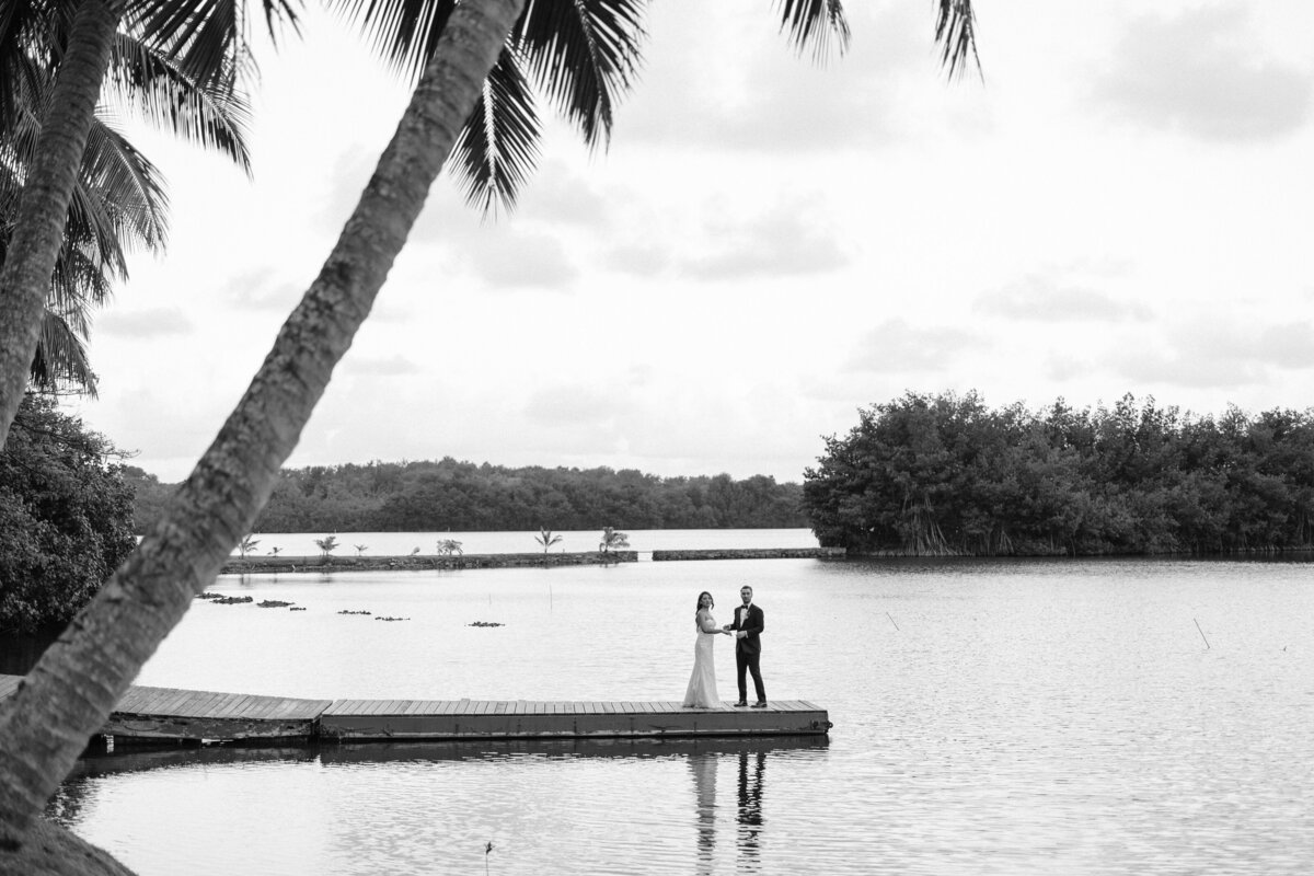 Wedding couple standing on dock in the middle of a  pond with palm tree in front of them