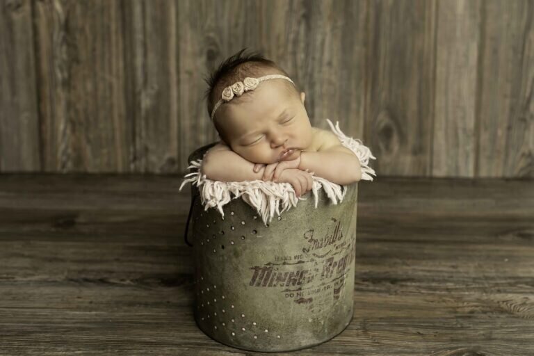 Newborn baby girl peacefully nestled in a rustic fishing bucket, wearing a delicate and pretty headband, creating a charming and whimsical scene