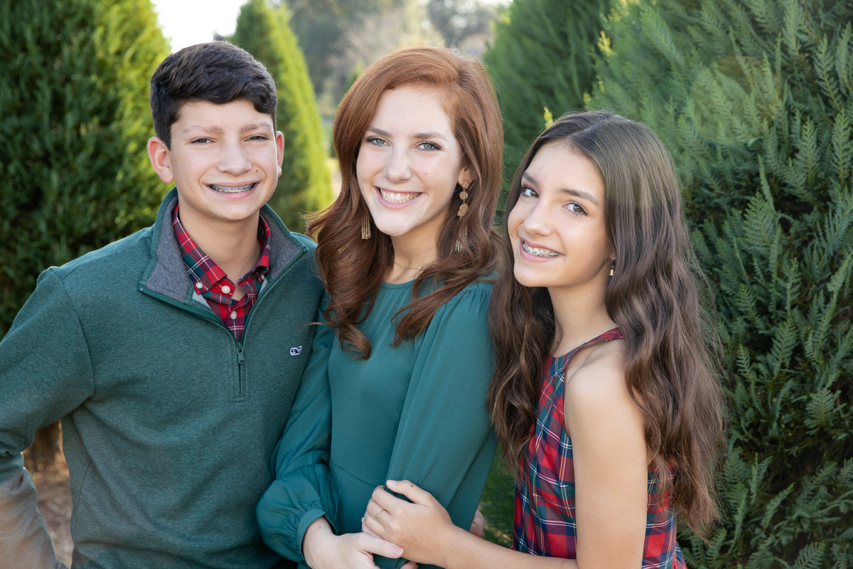 Tow girls and a boy smiling looking at the camera all wearing green tops standing in the middle of a tree farm