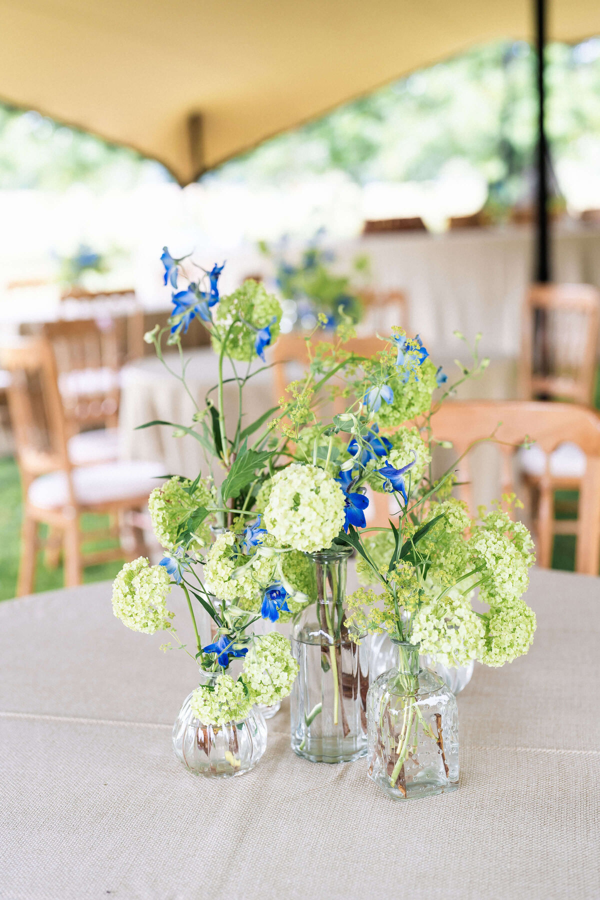 bud vases of green and blue flowers on a dining table outside under a canvas marquee for a birthday party afternoon tea at avington park
