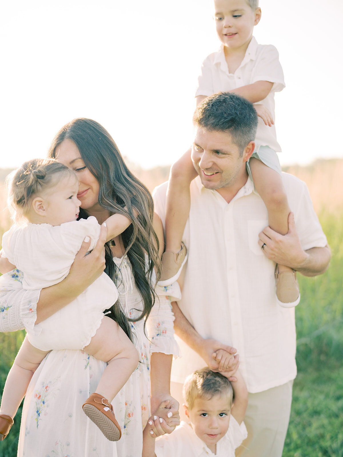 Mother holds toddler girl and leans into her while standing with her husband who has one son on his shoulders and one son in front of him during their Northern Virginia family session.