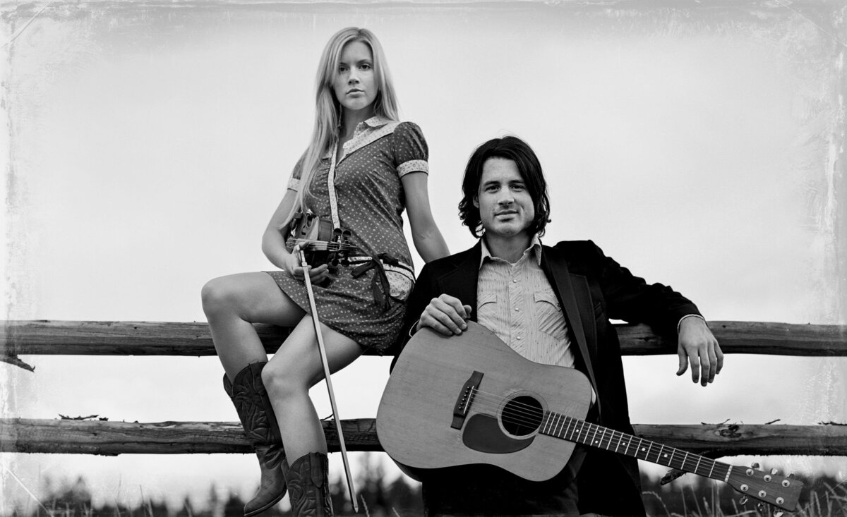 Musical duo portrait Dustin standing holding guitar Kendall sitting behind holding fiddle black and white
