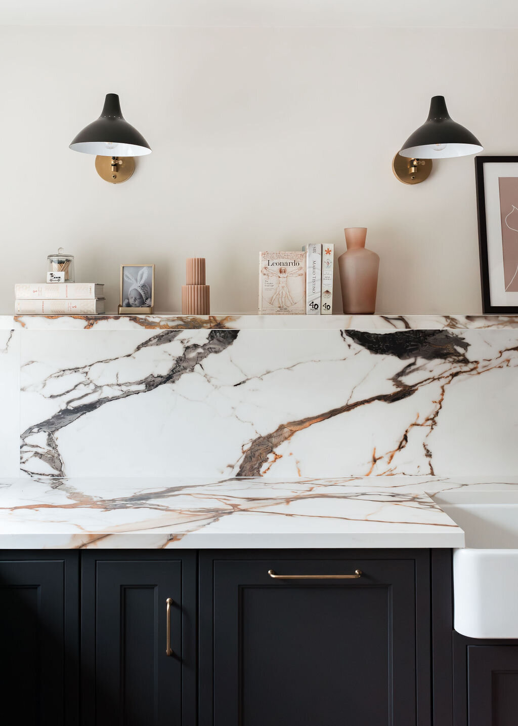 A modern kitchen featuring dark lower cabinets with a white marble countertop and backsplash. Wall-mounted black lights and neutral-toned decor add to the aesthetic