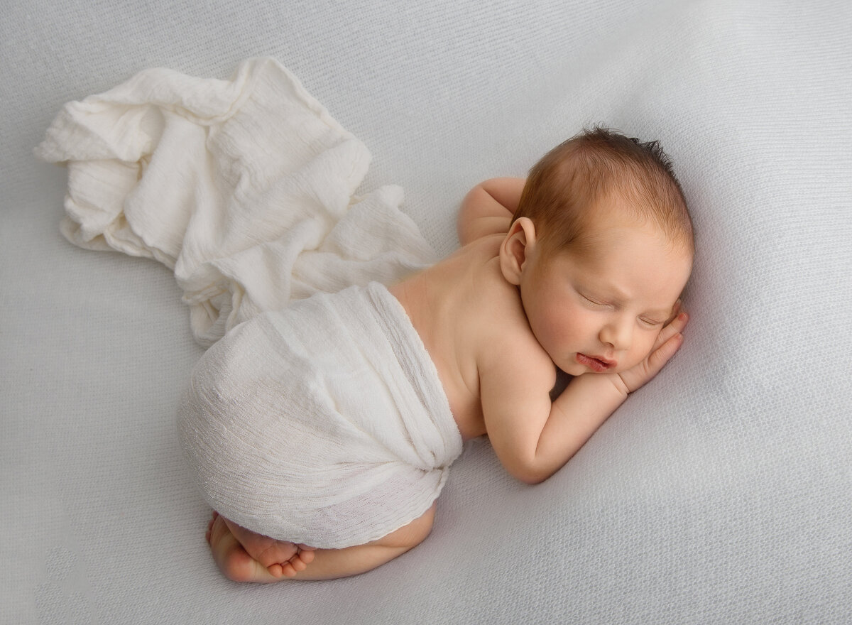 Baby is bare laying atop of his belly with his legs folded underneath him. Baby's hands are resting under his chin. Captured by top Brooklyn newborn photographer Chaya Bornstein