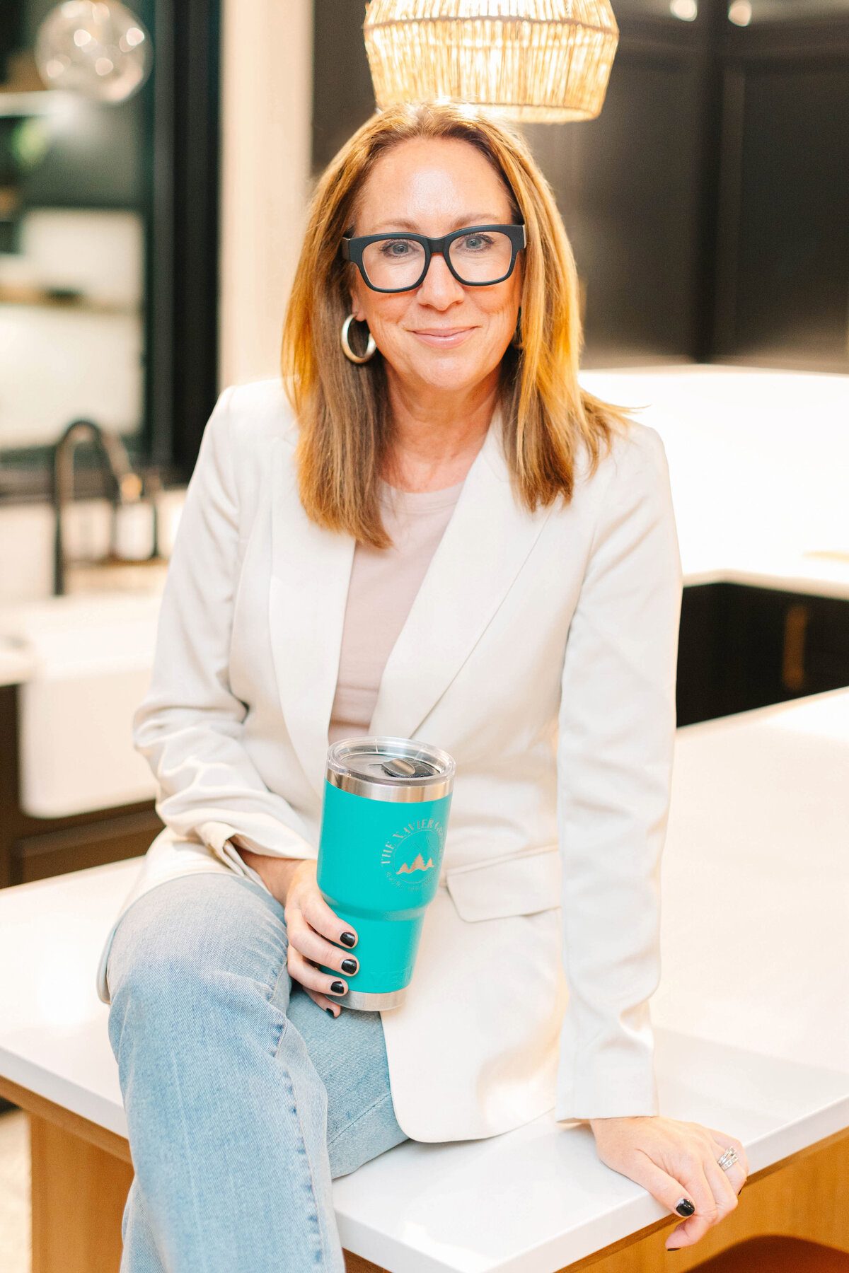 A stylish woman with shoulder-length blonde hair and black-framed glasses sits on a kitchen counter, holding a turquoise tumbler. She wears a white blazer, light jeans, and hoop earrings, with warm lighting and modern black cabinetry in the background