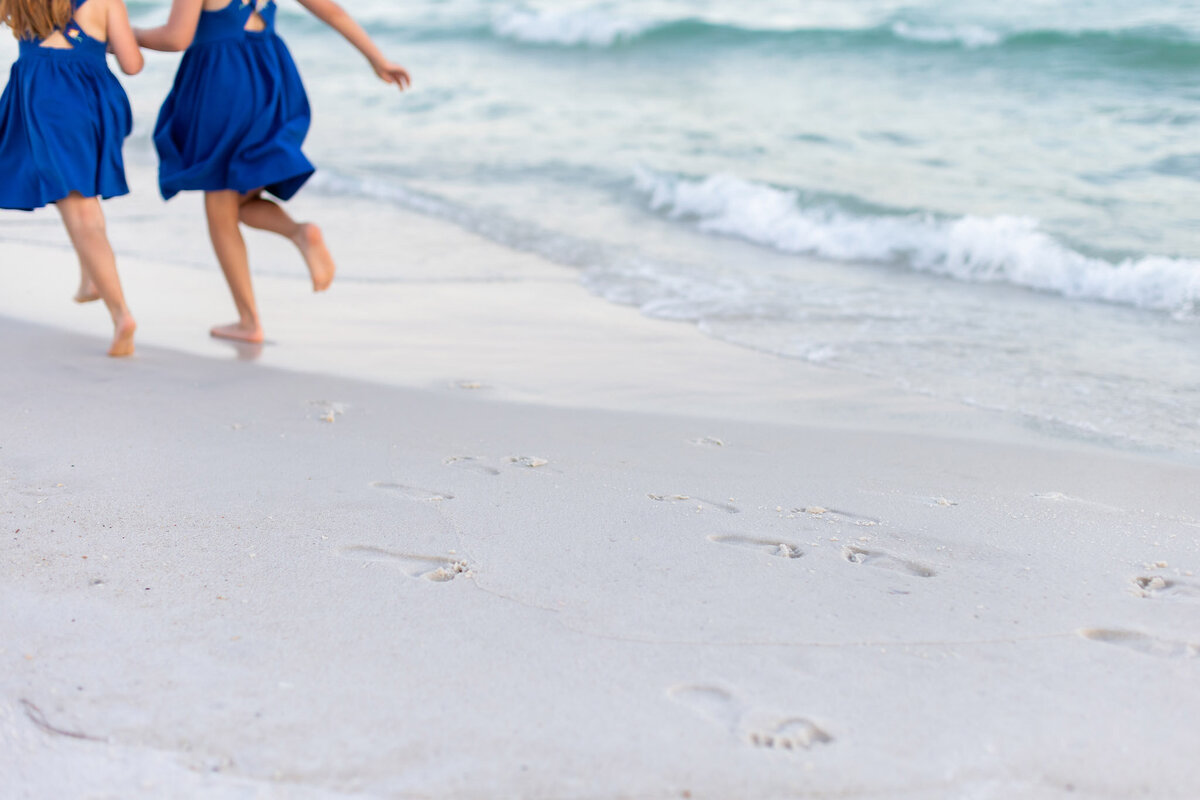 2 little girls wearing blue dresses are running away from the camera and only their legs and half of the dresses are captured in Rosemary Beach, FLorida.