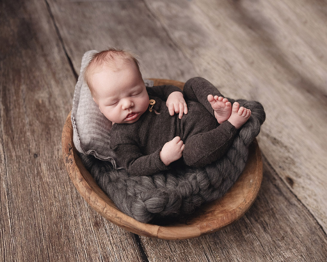 Baby boy sleeps in wood bowl with brown cozy sleeping outfit and newborn pillow.