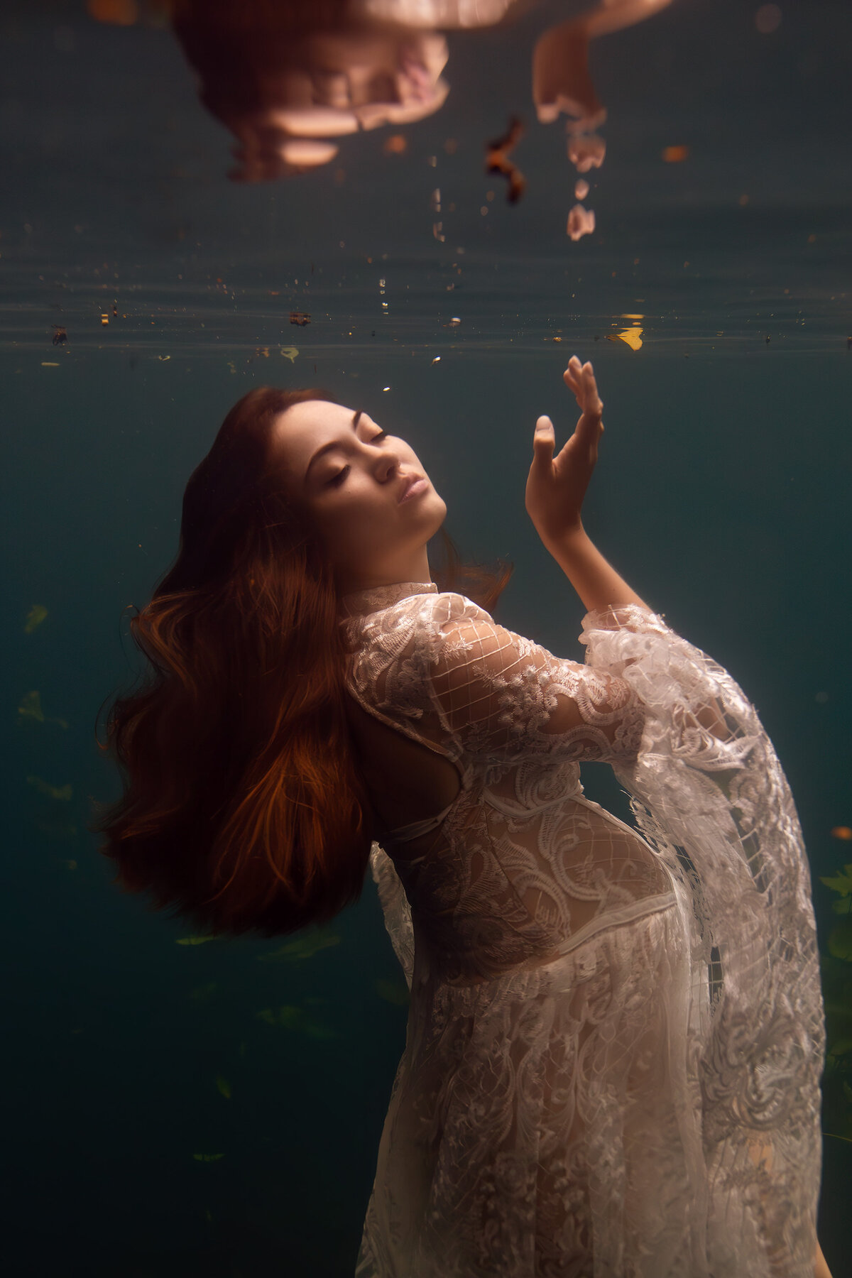 A serene underwater maternity photo featuring a woman gracefully floating in a delicate lace gown. Her face tilts upward towards the water's surface, where her reflection softly glows. The intricate lace pattern of her dress adds elegance to the tranquil scene, embodying the artistry of Dallas underwater maternity photographer Lindsay Walden.