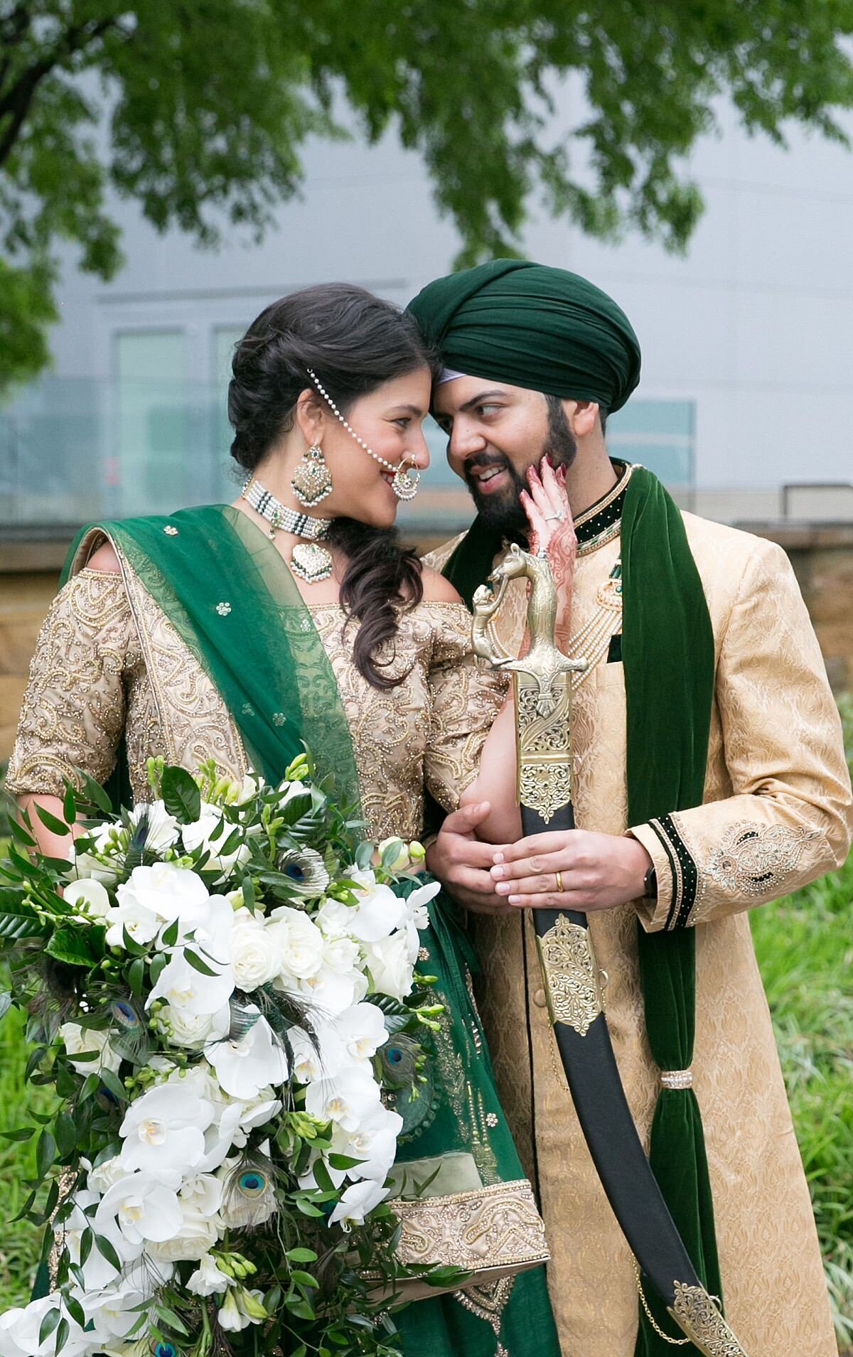 Sheikh groom wearing a green and gold sherwani and green turban holds a black sword with gold filagree ad he looks at the Indian bride. Hindu bride wearing a gold saree with a green dupatta holds a cascading bouquet of white orchids as she looks into the groom's eyes in Nashville, TN