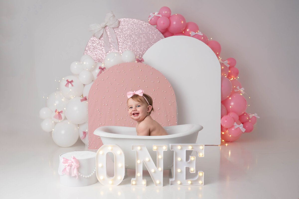 Smiling baby leaning forward in a mini tub, with LED letters in front and a small cake prop nearby.