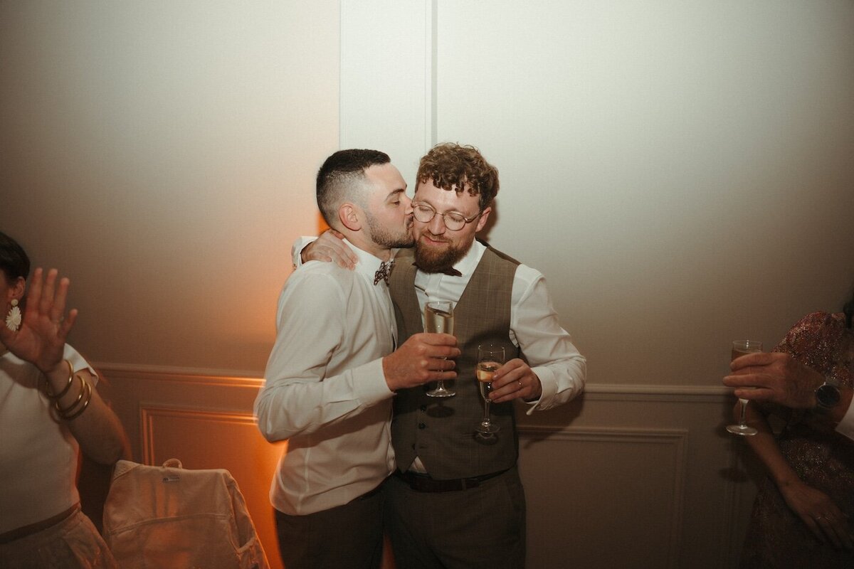 Un invité en nœud papillon embrasse le marié sur la joue, un verre de champagne à la main. Mariage par Kamescop, photographe et vidéaste vintage.