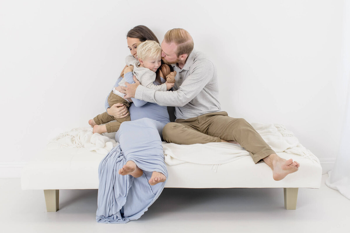 A family sits on a white bed against a white background. A woman in a blue dress holds a young child, while a man in a gray shirt and beige pants embraces them both. The scene conveys warmth and affection.
