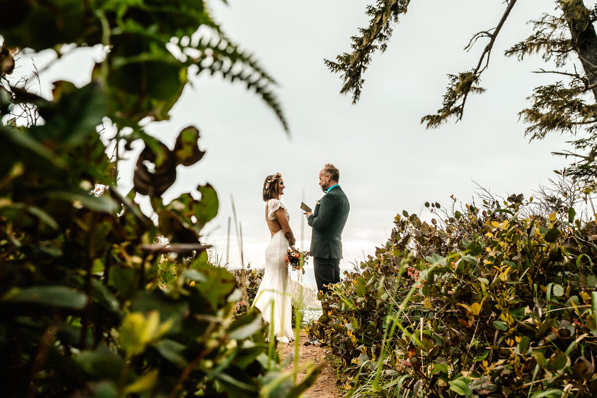 during their oregon coast elopement, a bride and groom exchange vows, surrounded by lush coastal greenery.