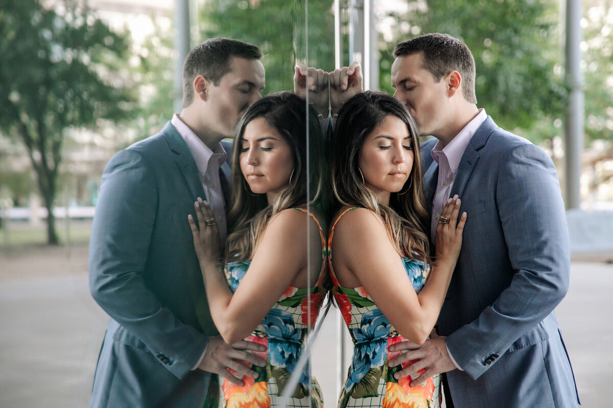 Engaged couple in Dallas Arts District at the Winspear Opera, she leaning on a glass wall while he is kissing her on the temple, their reflection visible in the glass