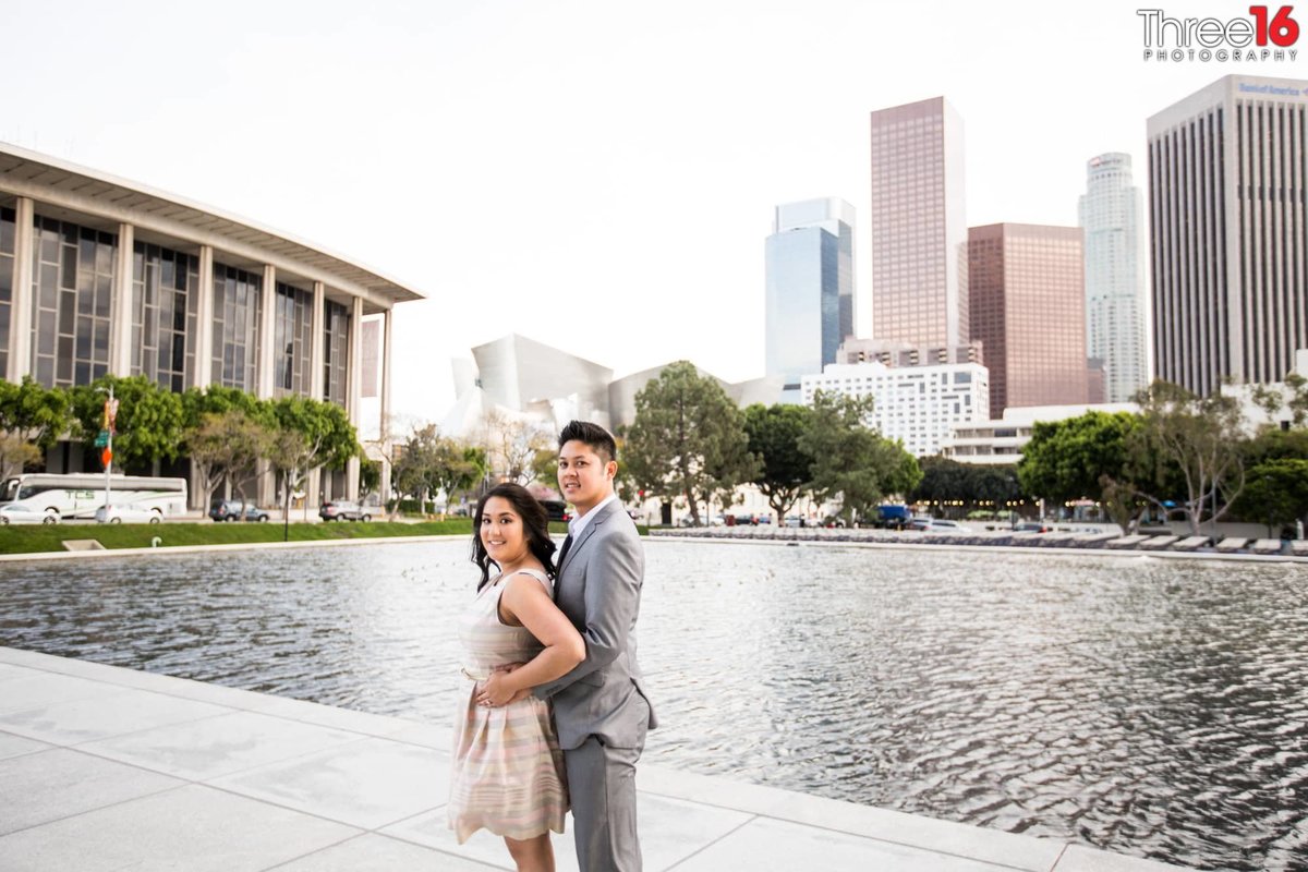 Groom to be holds his Bride from behind as they pose in front of Downtown LA fountain