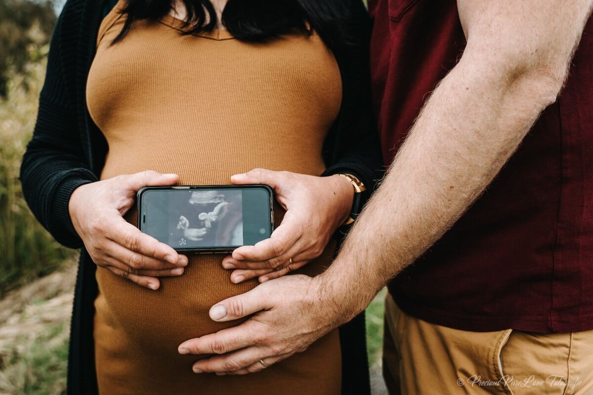 Outdoor maternity photo of a pregnant woman in a mustard dress holding a smartphone displaying her baby’s ultrasound image over her baby bump, with her partner’s hands lovingly resting on her belly.