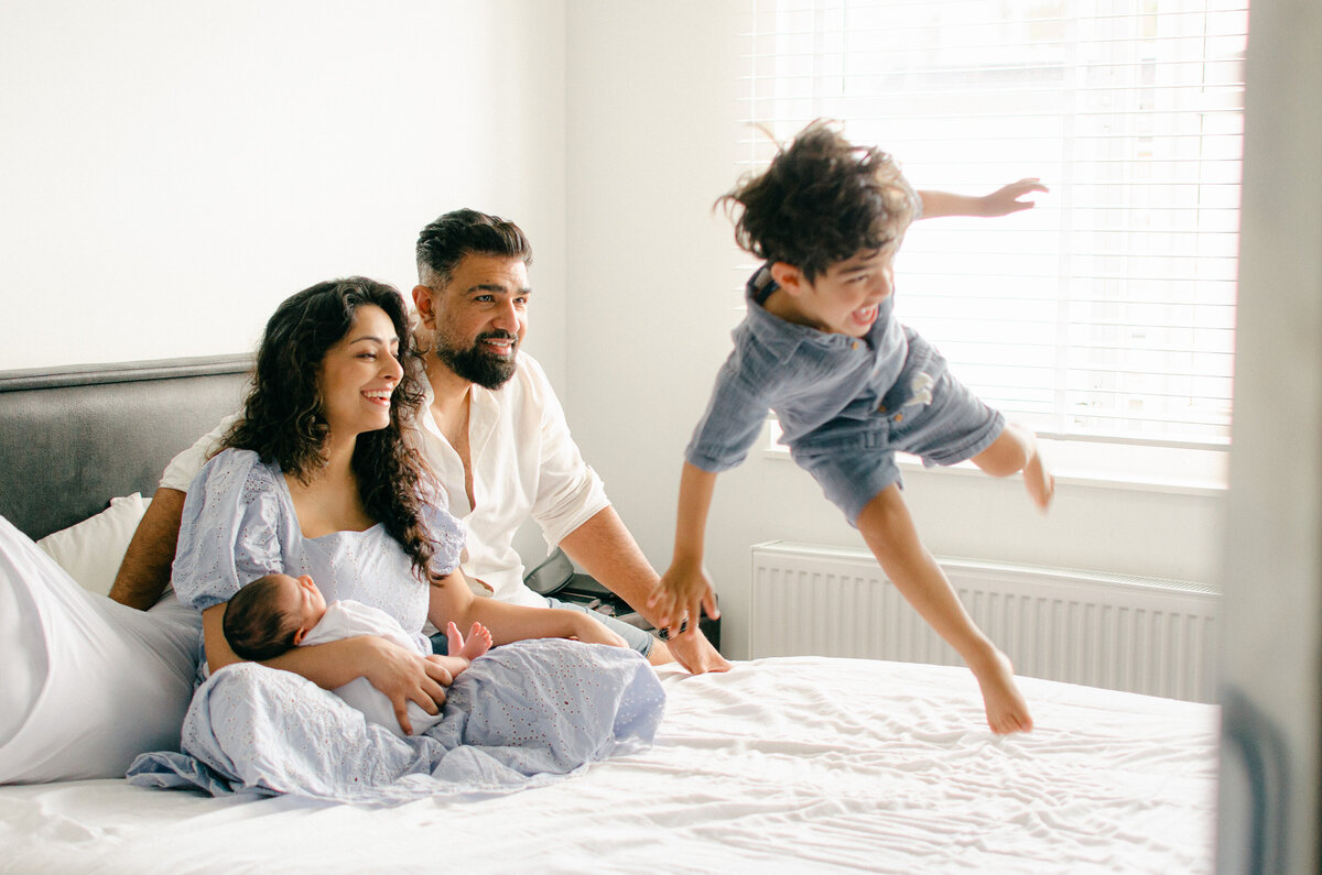 Husband and wife hold their newborn while their oldest jumps on the bed. 