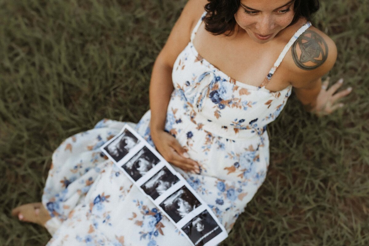Woman in floral dress sitting on grass, holding ultrasound images on her lap, and touching her belly.