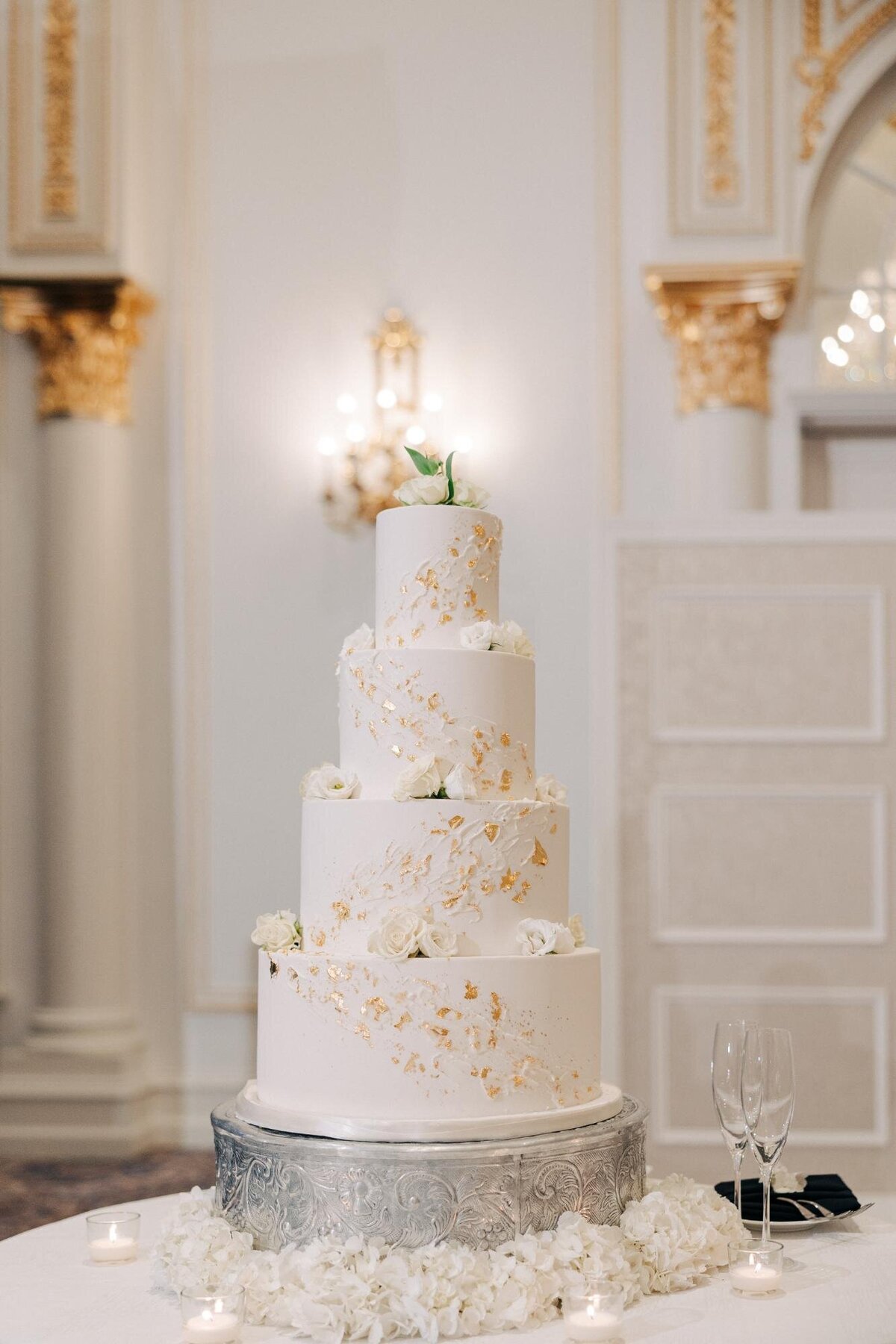 Elegant multi-tiered wedding cake with gold leaf accents on a silver stand, surrounded by candles and white flowers in a luxurious interior.