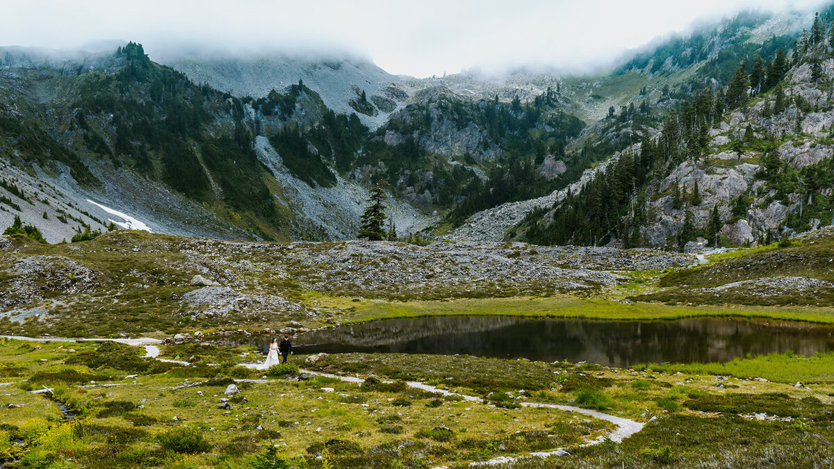 a landscape shot of this mountain and alpine lake scene. The adventure elopement photographer lines a couple in wedding attire up on the first third. They look small in the massive landscape as they walk a long the  dirt path, holding hands