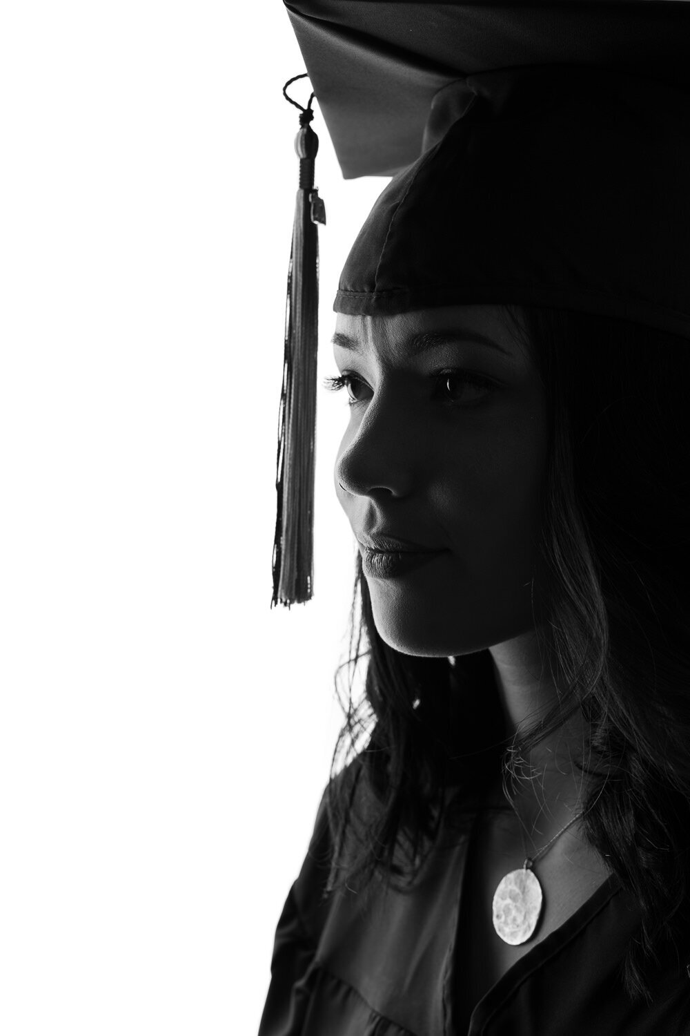 Editorial graduation portrait featuring a high-contrast silhouetted face of a senior wearing her cap and gown, photographed against a white studio background.