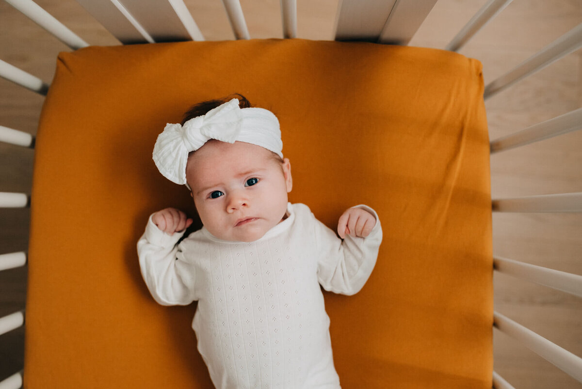 A baby girl lays against a burnt orange colored sheet in her crib while wearing a white onesie and white bow, she is looking at the camera