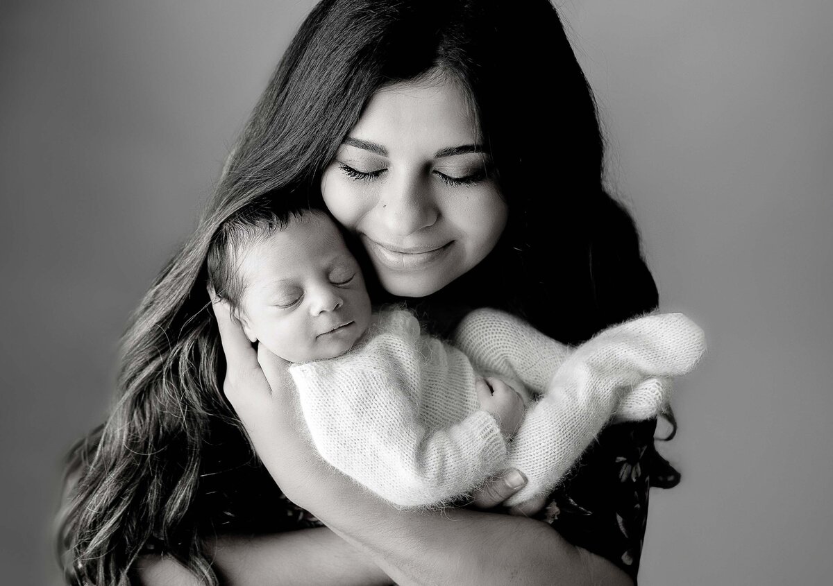 Newborn studio photo shoot with top Philadelphia and Main Line newborn photographer. Mom is cradling baby in her arms against her upper chest. Her eyes are closed and resting her cheek against baby's temple. Baby's eyes are close and sleeping peacefully.