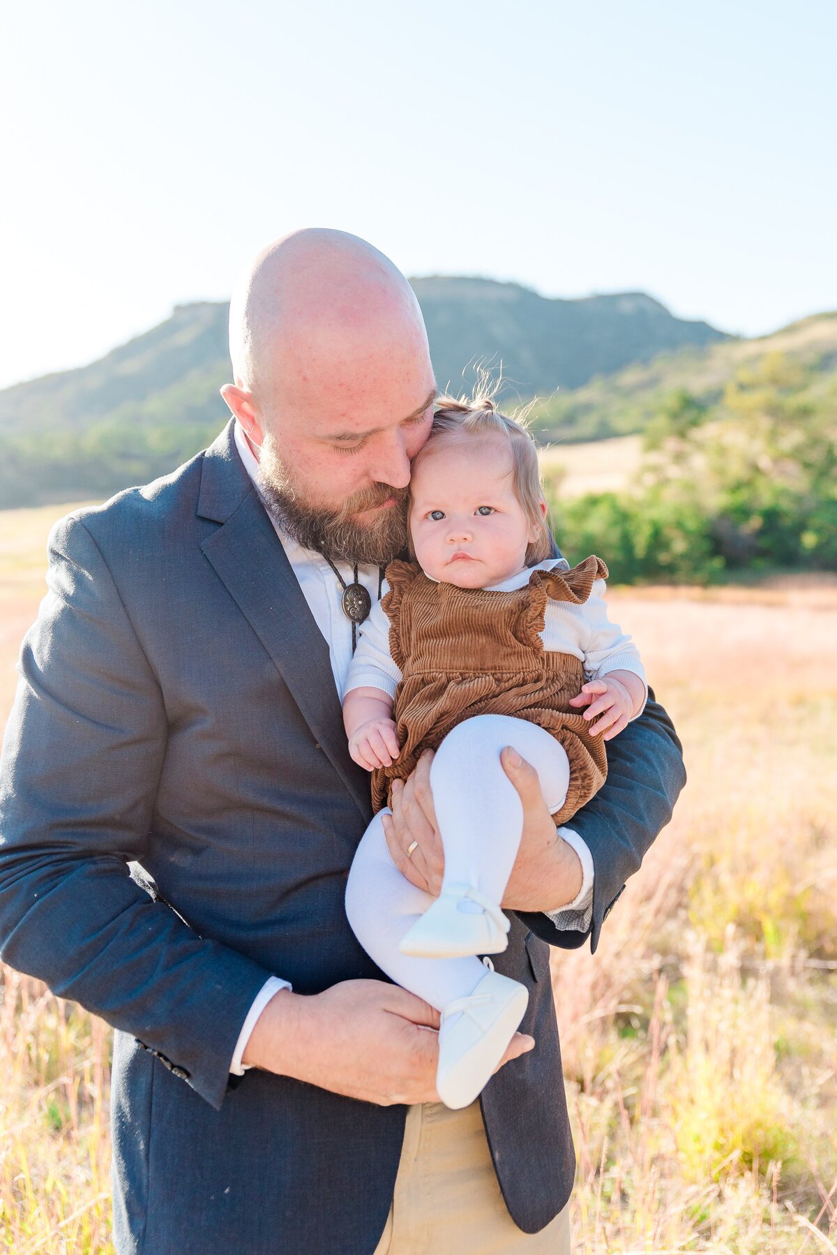 A dad holds his infant daughter in a brown overall dress and looks down at her captured by denver family photographer