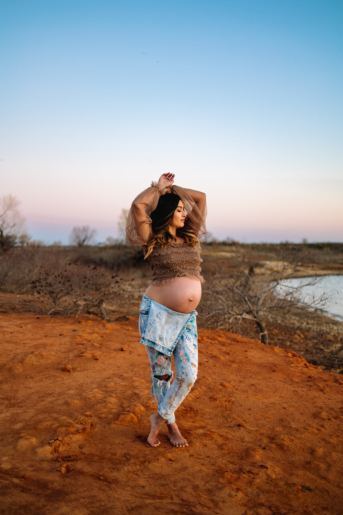 Maternity photography featuring a pregnant woman posing in the sand. She is dressed in blue jeans and a short brown shirt, with her arms raised above her head and eyes closed.