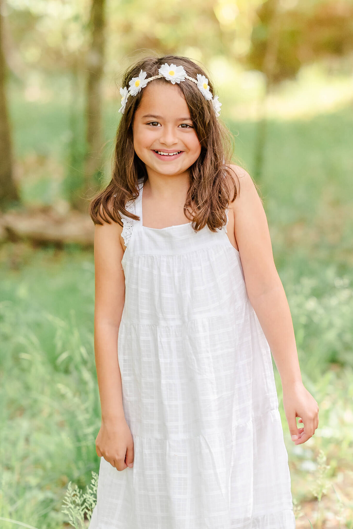 A young girl wearing a white dress and flower crown smiles for the camera at a park in Virginia Beach.