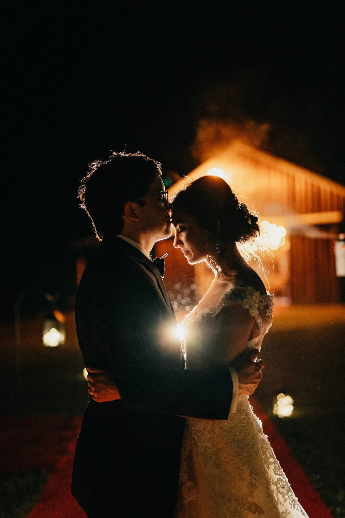 A night-time backlit photo of a bride and groom embracing, with a warm glow surrounding them