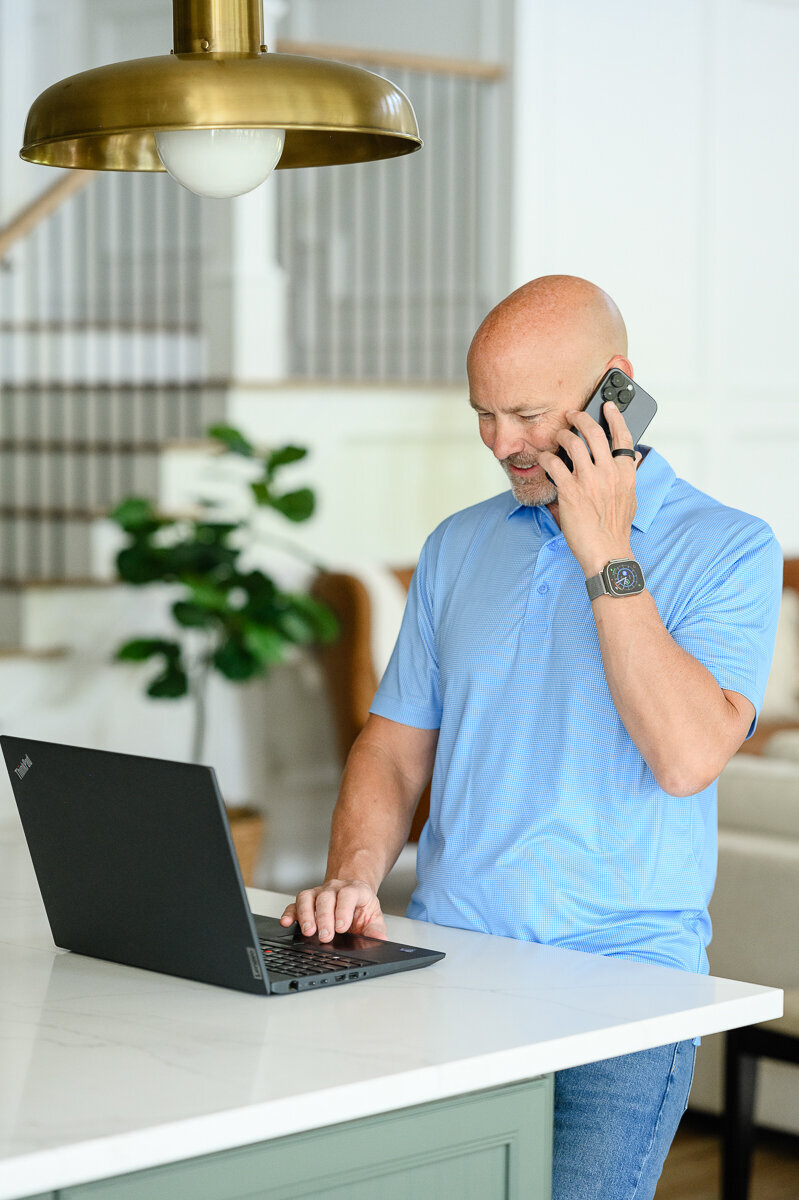 A mortgage broker stands at a kitchen counter on his computer smiling at a brand photographer in Denver for a branding photography photo shoot and brand photos.
