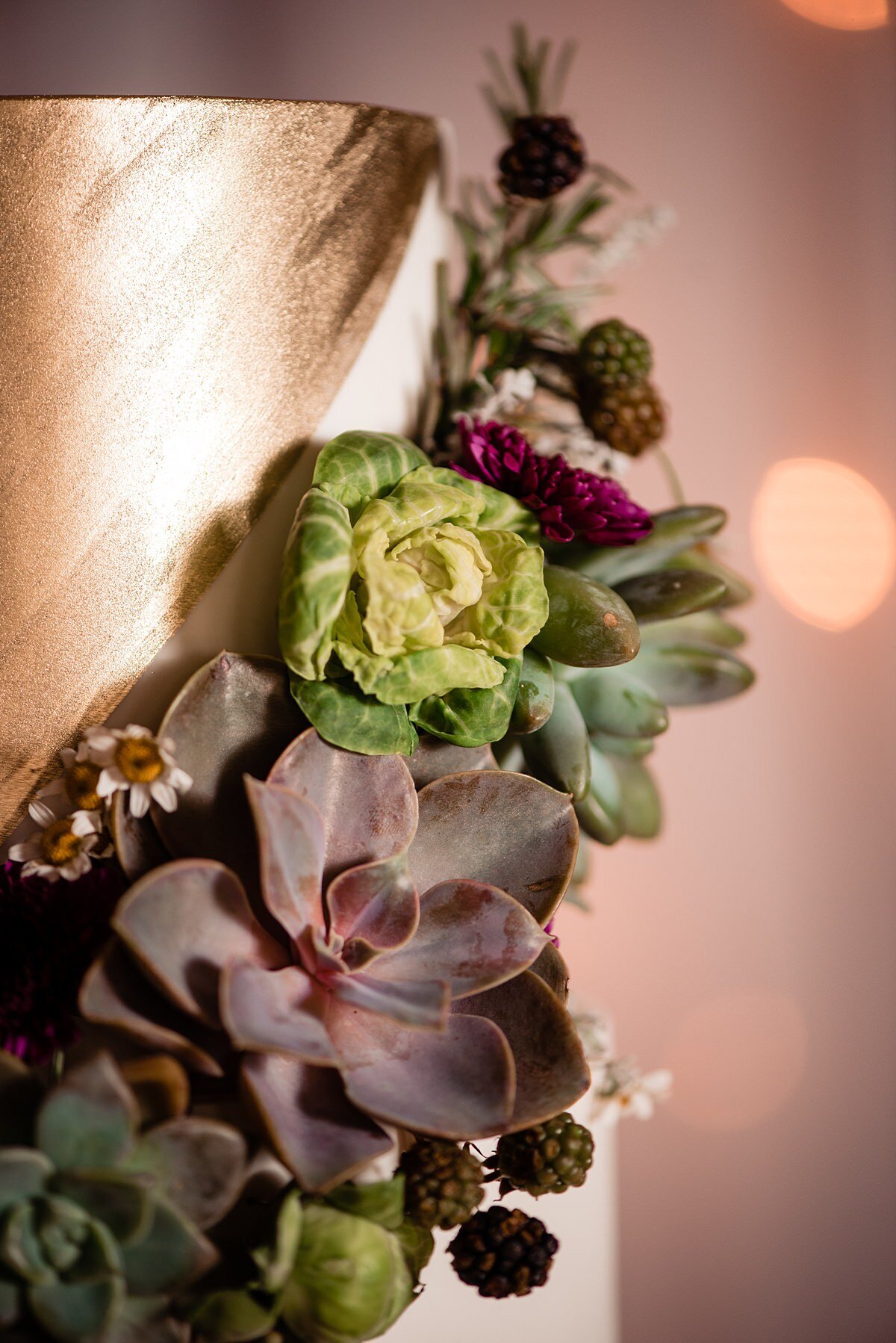 A gold and white wedding cake decorated with a garland of succulents, blackberries and brussel sprouts at City Winery Nashville