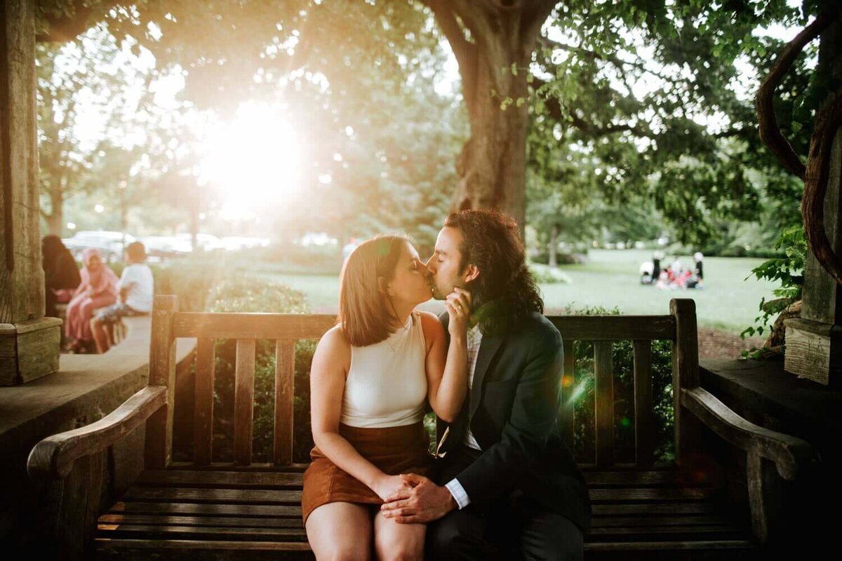 Couple sharing a kiss sitting on a bench in a park backlit by the setting sun in Philadelphia,