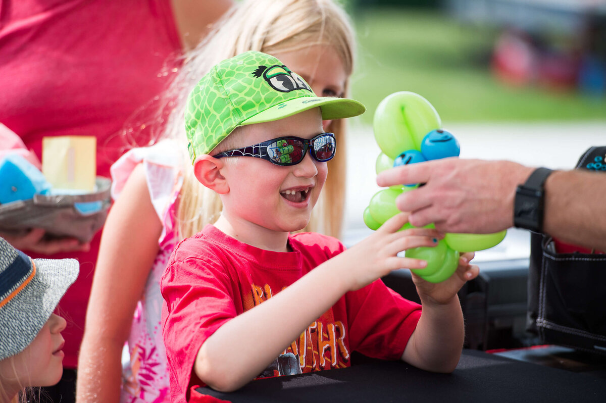 a little boy in a red t-shirt showing excitement on his face and smiling as he receives a balloon animal as part of a corporate children's event.  Captured by Ottawa Event Photographer JEMMAN Photography COMMERCIAL