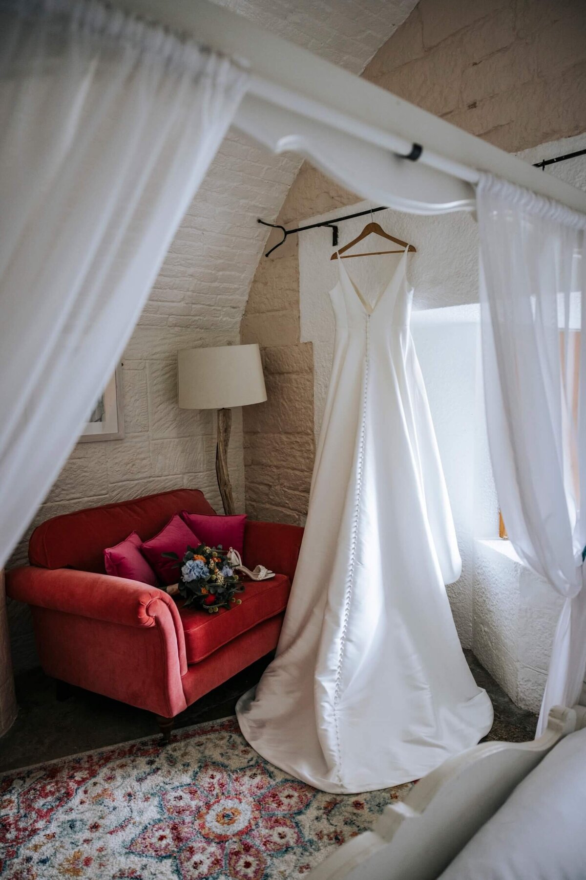 A bed room with a red chair and a canopy with a wedding dress hanging in front of the window