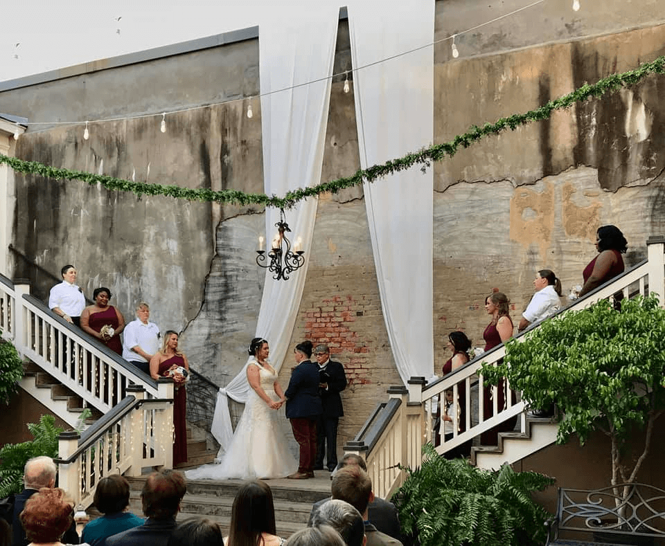A wedding ceremony in an outdoor courtyard with two brides standing at the altar between two staircases, surrounded by wedding parties dressed in white and burgundy. Guests are seated in the foreground.