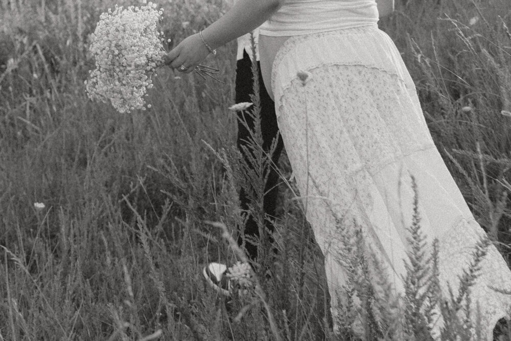 A cinematic photo of a couple walking through a grassy field. The man is leading his pregnant wife while she carries a bouquet of flowers in Johnson City, TN. 